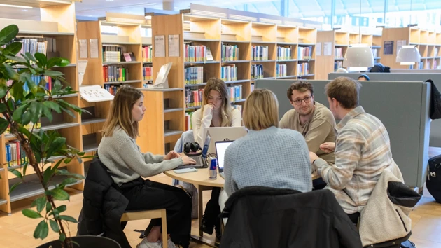 Tre personer som sitter vid ett bord i biblioteket och pluggar. Runt bordet syns flera bokhyllor med böcker.