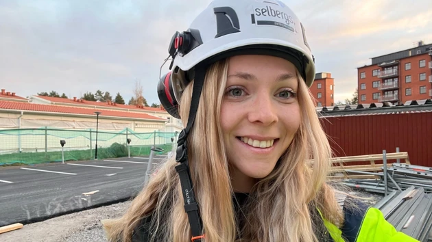 Porträttbild på Wilma Edin i vit bygghjällm, husbygge i bakgrunden